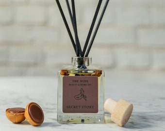 Peut inclure: Un diffuseur de parfum en verre transparent avec des roseaux noirs et un bouchon en bois. La bouteille porte une étiquette sur laquelle est inscrit "The Nine Beauty & Home Spa Secret Story". Il y a deux petits bonbons au chocolat au premier plan.