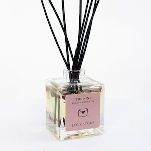 Puede incluir: Un difusor de vidrio transparente con varillas negras. La botella contiene un líquido amarillo y una etiqueta rosa que dice "LOVE STORY". La etiqueta también tiene un gráfico de corazón y sobre. El producto es de "THE NINE BEAUTY & HOME SPA".