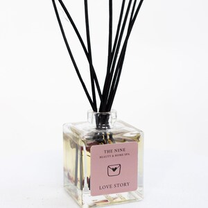 Könnte beinhalten: Eine klare Glasflasche mit schwarzen Diffusorstäbchen und einem rosa Etikett mit der Aufschrift "THE NINE BEAUTY & HOME SPA" und "LOVE STORY".