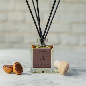 Könnte beinhalten: Ein klarer Glasdiffusor mit schwarzen Schilfrohren und einem Holzdeckel. Die Flasche hat ein Etikett mit der Aufschrift "The Nine Beauty & Home Spa Secret Story". Im Vordergrund befinden sich zwei kleine Schokoladenbonbons.