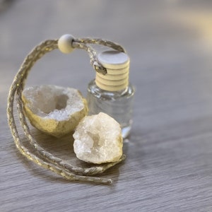 Può includere: Una bottiglia di vetro trasparente con un tappo di legno bianco e un cordoncino intrecciato beige. La bottiglia è appoggiata su una superficie di legno con due geodi di cristallo bianco.
