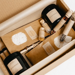 K&ouml;nnte beinhalten: Ein brauner Karton gef&uuml;llt mit verschiedenen Artikeln, darunter schwarze Gl&auml;ser mit der Aufschrift "MOOD KERZE", kleine Glasflaschen und braune Papierverpackungen. Die Box enth&auml;lt Artikel f&uuml;r ein Spa- oder Beauty-Set.