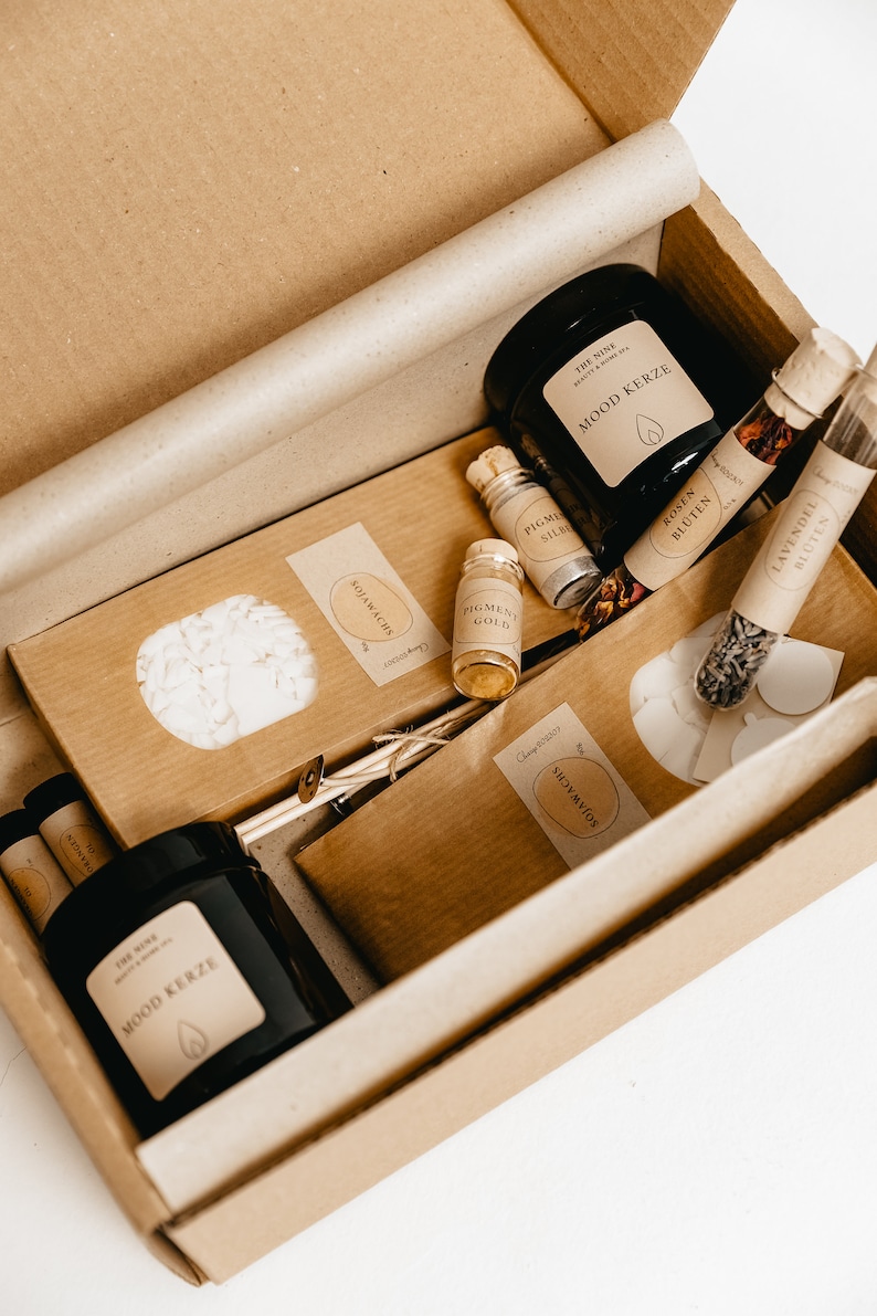 K&ouml;nnte beinhalten: Ein brauner Karton gef&uuml;llt mit verschiedenen Artikeln, darunter schwarze Gl&auml;ser mit der Aufschrift "MOOD KERZE", kleine Glasflaschen und braune Papierverpackungen. Die Box enth&auml;lt Artikel f&uuml;r ein Spa- oder Beauty-Set.