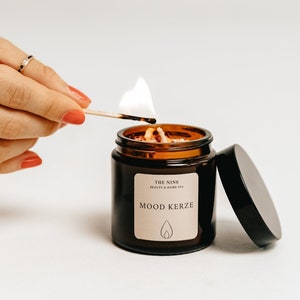K&ouml;nnte beinhalten: Eine brennende braune Glas-Kerze mit dem Text "MOOD KERZE" auf einem Etikett. Die Kerze wird mit einem Streichholz angez&uuml;ndet. Die Kerze ist von "THE NINE BEAUTY & HOME SPA".