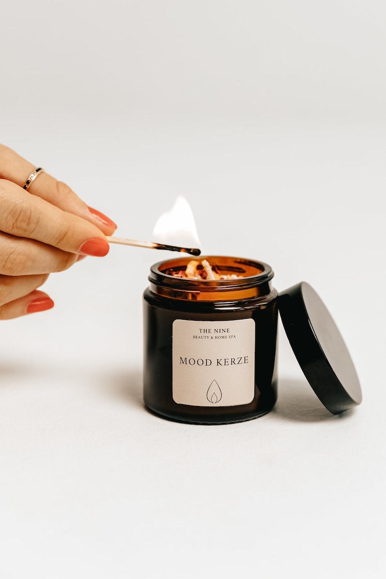 K&ouml;nnte beinhalten: Eine brennende braune Glas-Kerze mit dem Text "MOOD KERZE" auf einem Etikett. Die Kerze wird mit einem Streichholz angez&uuml;ndet. Die Kerze ist von "THE NINE BEAUTY & HOME SPA".