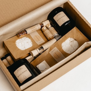K&ouml;nnte beinhalten: Eine Geschenkbox mit Aromatherapie-Artikeln. Die Box enth&auml;lt eine schwarze Kerze mit der Aufschrift "MOOD KERZE", kleine Glasfl&auml;schchen und P&auml;ckchen mit Aufschriften wie "ROSEN BLUTEN" und "SOJAWACHS". Die Artikel sind in einer braunen Pappschachtel angeordnet.
