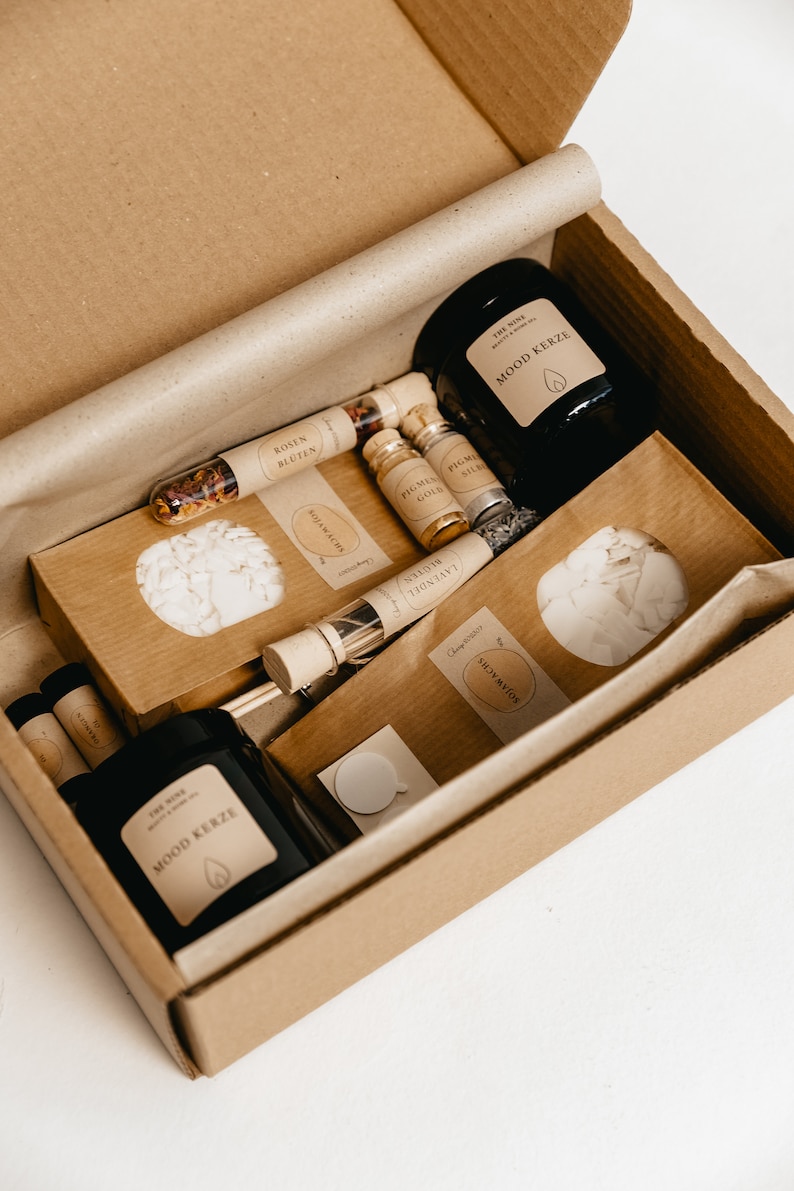 K&ouml;nnte beinhalten: Eine Geschenkbox mit Aromatherapie-Artikeln. Die Box enth&auml;lt eine schwarze Kerze mit der Aufschrift "MOOD KERZE", kleine Glasfl&auml;schchen und P&auml;ckchen mit Aufschriften wie "ROSEN BLUTEN" und "SOJAWACHS". Die Artikel sind in einer braunen Pappschachtel angeordnet.