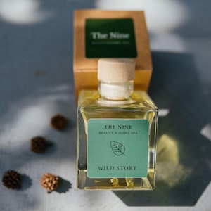 Könnte beinhalten: Eine klare Glasflasche mit einem Holzverschluss und einem grünen Etikett mit der Aufschrift "THE NINE BEAUTY & HOME SPA WILD STORY".