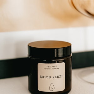 K&ouml;nnte beinhalten: Ein dunkelbraunes Glas mit einem Holzdeckel. Das Etikett lautet "MOOD KERZE" mit einem Flammensymbol. Das Glas stammt von "THE NINE BEAUTY & HOME SPA". Der Hintergrund umfasst ein verschwommenes Bild und ein goldfarbenes Glas.