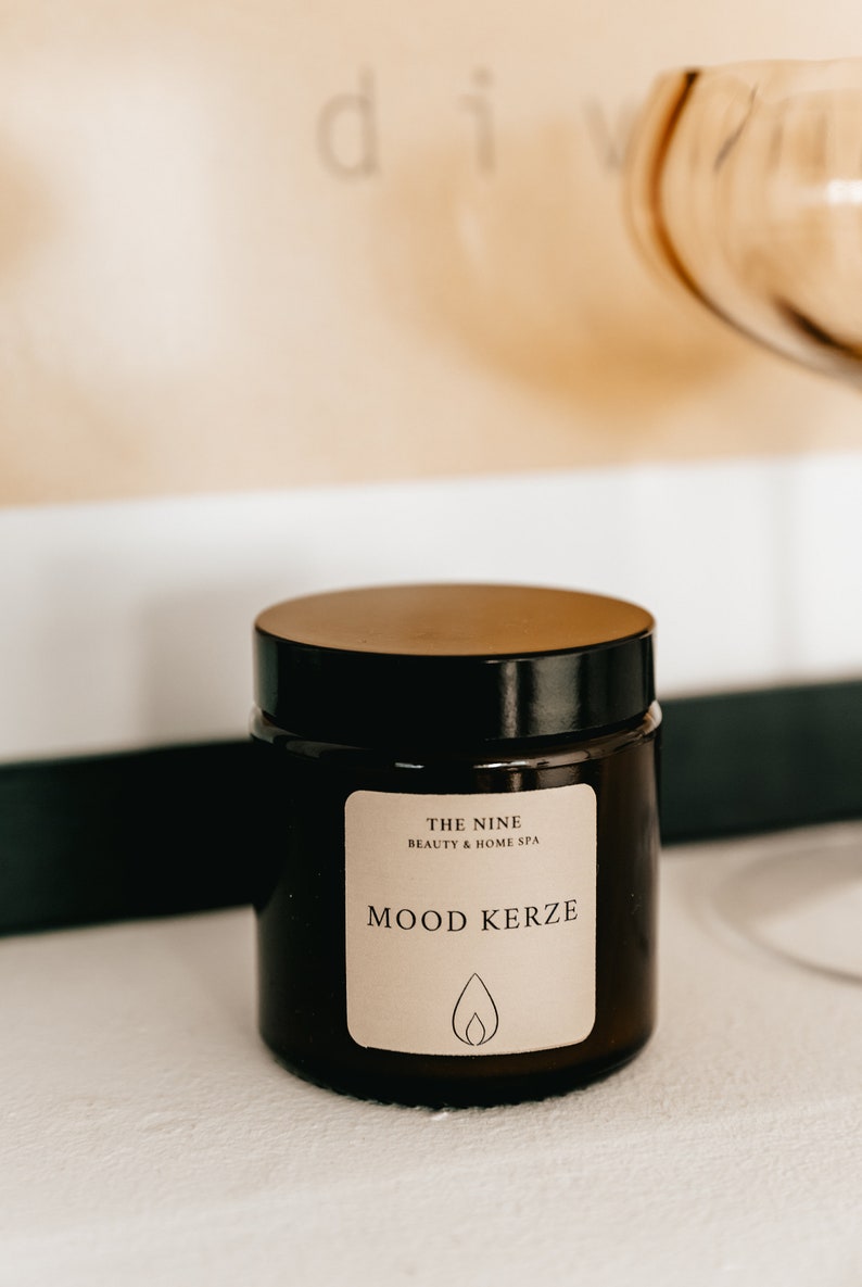 K&ouml;nnte beinhalten: Ein dunkelbraunes Glas mit einem Holzdeckel. Das Etikett lautet "MOOD KERZE" mit einem Flammensymbol. Das Glas stammt von "THE NINE BEAUTY & HOME SPA". Der Hintergrund umfasst ein verschwommenes Bild und ein goldfarbenes Glas.