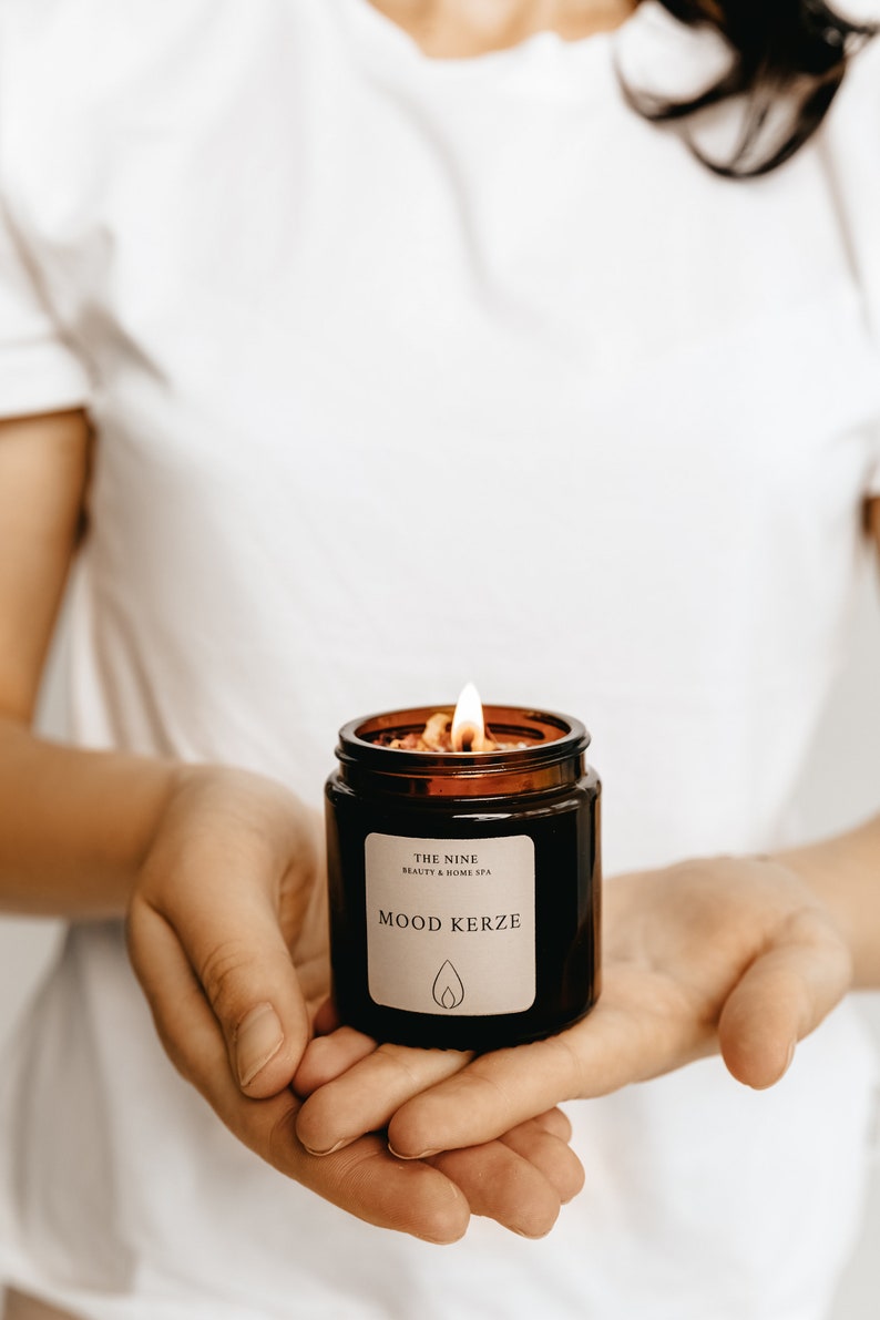 K&ouml;nnte beinhalten: Eine brennende, braune Glas-Kerze mit dem Text "MOOD KERZE" auf einem wei&szlig;en Etikett. Die Kerze ist mit Wachs und kleinen St&uuml;cken getrockneter Blumen gef&uuml;llt. Die Kerze wird in offenen H&auml;nden gehalten.