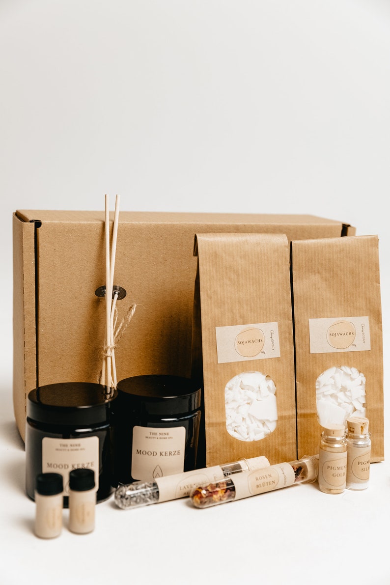 K&ouml;nnte beinhalten: Ein Geschenkset mit einer braunen Pappschachtel, zwei schwarzen Gl&auml;sern mit der Aufschrift "Mood Kerze" und zwei braunen Papiert&uuml;ten mit dem Wort "Sojawachs". Ebenfalls enthalten sind kleine Glasfl&auml;schchen und ein B&uuml;ndel Holzst&auml;bchen.