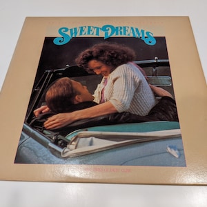 May include: A vinyl record album cover featuring a couple in a convertible car. The album cover is titled "Sweet Dreams" and features the name "Patsy Cline" in smaller font. The album cover is beige with a photo of a couple in a convertible car.