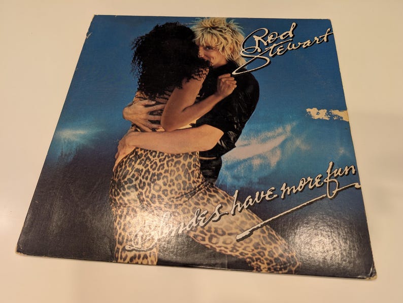 Puede incluir: Portada del &aacute;lbum "Blondes Have More Fun" de Rod Stewart. La portada presenta a un hombre y una mujer abraz&aacute;ndose, con el texto "Rod Stewart" y "Blondes Have More Fun" en una fuente estilizada sobre un fondo de cielo azul.