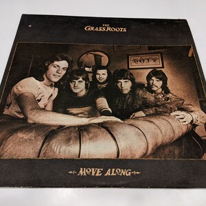 May include: The Grass Roots album cover, featuring a sepia-toned photo of the band members sitting on a couch. The album title "Move Along" is printed in gold lettering on a black background.