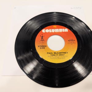 May include: A black vinyl record with a vibrant orange and yellow label. The label features the text "Columbia" and "Paul McCartney Take It Away". The record is a 45 RPM single.