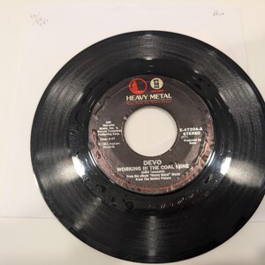 May include: A black vinyl record with a white center label featuring the text "HEAVY METAL" and the band name "DEVO". The record is titled "WORKING IN THE COAL MINE" and is from the movie "Heavy Metal".