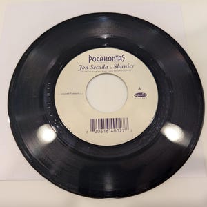 May include: A black vinyl record with a white label featuring the title "Pocahontas" and the names "Jon Secada & Shanice." The label also includes a barcode and the text "English Version."