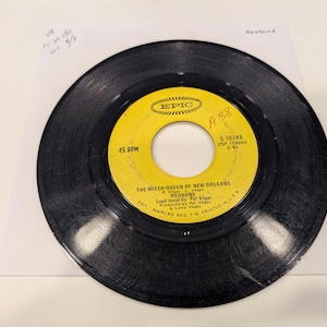May include: A black vinyl record with a yellow label featuring the text "THE WITCH QUEEN OF NEW ORLEANS" by Redbone. The label also includes the Epic Records logo and the speed of 45 RPM.