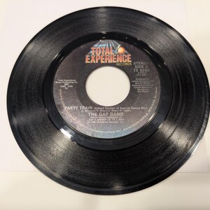 May include: A black vinyl record with the label "Total Experience Records" and the song title "Party Train" by The Gap Band. The record is centered on a white surface, showcasing its circular shape and grooves.