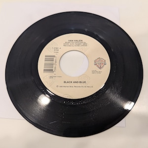 May include: A black vinyl record with a white label featuring the text "VAN HALEN" and "BLACK AND BLUE." The record is a 7-inch single, with the Warner Bros. Records logo visible. The record is on a white surface.