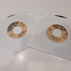 May include: Two vinyl records in white sleeves. One record label reads "Maniac (Vocal) Michael Sembello". The other record label reads "Flashdance... What a Feeling Irene Cara". Both records are 45 RPM.