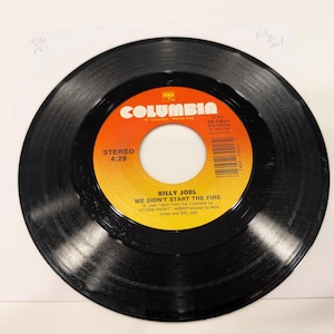 May include: A black vinyl record with a Columbia label featuring orange and yellow gradients. The label displays the text "BILLY JOEL WE DIDN'T START THE FIRE" and "STEREO 4:29". The record is on a white paper sleeve.