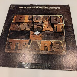 May include: Vintage vinyl record album cover for Blood, Sweat & Tears' Greatest Hits. The album cover features the band's name in large, three-dimensional wooden block letters. The background is a dark, textured surface.