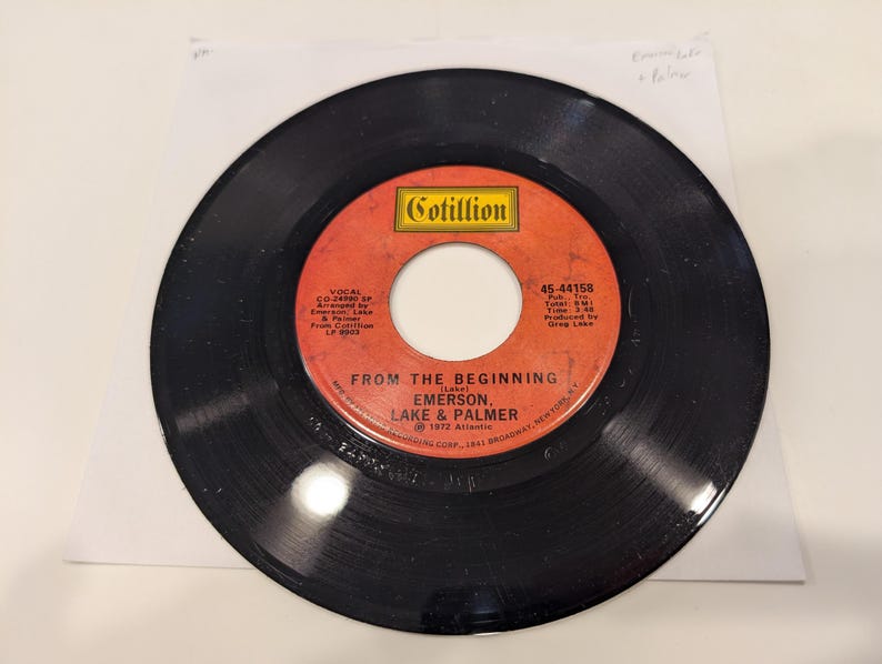 May include: A black vinyl record with a red and orange label. The label reads "From The Beginning" by Emerson, Lake & Palmer. The record is a 45 RPM single from Cotillion Records.