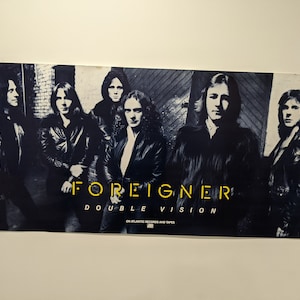 Può includere: Un poster vintage per l'album "Double Vision" della band Foreigner. Il poster presenta una foto in bianco e nero dei membri della band davanti a un muro di mattoni. Il nome della band e il titolo dell'album sono stampati in lettere gialle.