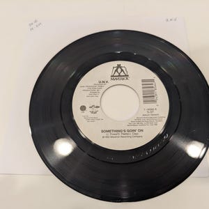 May include: A black vinyl record with a white label featuring the text "SOMETHING'S GOIN' ON" by U.N.V. The record is centered on a white background, with the Maverick logo visible.
