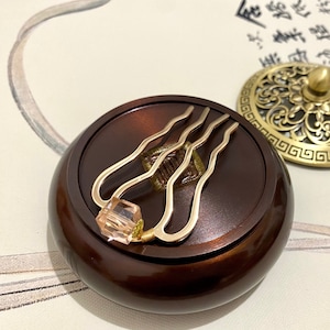 Pode incluir: Um queimador de incenso de madeira marrom com uma tampa dourada e um pente decorativo dourado no topo. A tampa tem um design floral e uma pequena maçaneta no topo.