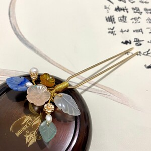 May include: A gold hair stick with a decorative floral design. The design features a pink flower, blue and green leaves, and yellow and white beads.