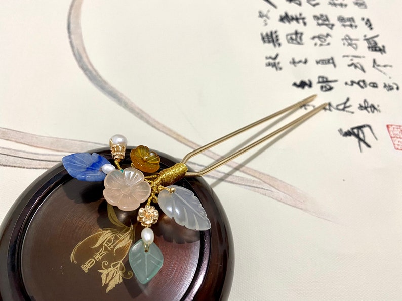 May include: A gold hair stick with a decorative floral design. The design features a pink flower, blue and green leaves, and yellow and white beads.