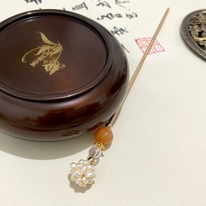 May include: A dark brown wooden incense burner with a gold inscription. A gold incense stick with a brown bead and a cluster of white pearls hangs from the burner.
