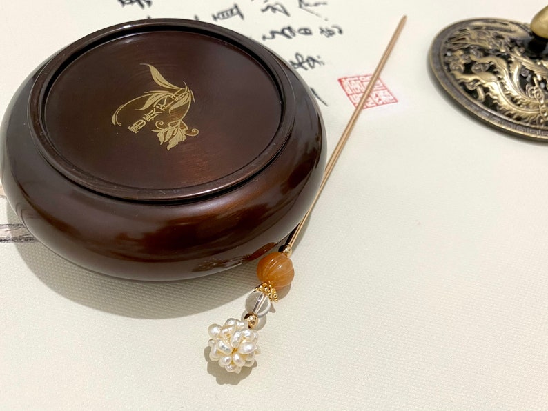 May include: A dark brown wooden incense burner with a gold inscription. A gold incense stick with a brown bead and a cluster of white pearls hangs from the burner.