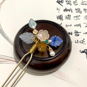 May include: A gold hair stick with a floral design made of jade, pearl, and other stones. The stick is resting on a wooden base.