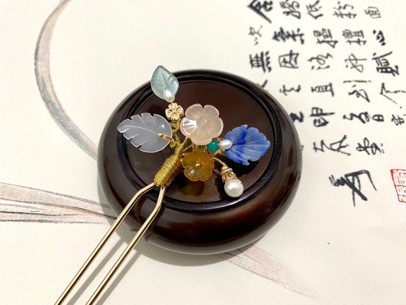 May include: A gold hair stick with a floral design made of jade, pearl, and other stones. The stick is resting on a wooden base.