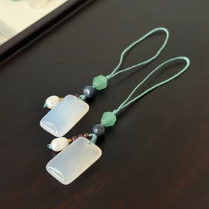 May include: Two rectangular, pale jade pendants with light blue cords. Each pendant is adorned with green, blue, and purple beads, and a white pearl. The pendants are displayed on a dark brown surface.