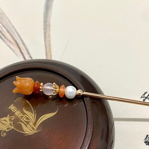 K&ouml;nnte beinhalten: Ein goldener Haarstecker mit einer geschnitzten orangefarbenen Blume und Perlen. Der Stecker liegt auf einer braunen Holzoberfl&auml;che mit einem goldenen Blumendesign.