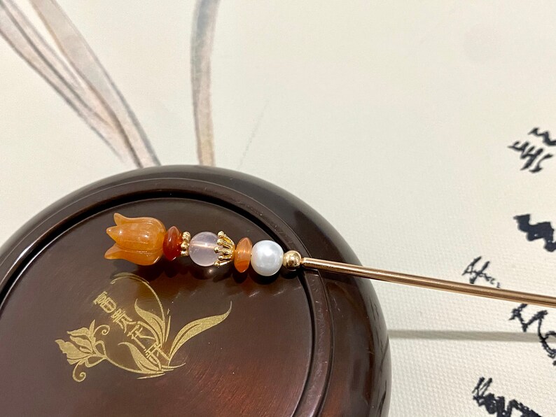 K&ouml;nnte beinhalten: Ein goldener Haarstecker mit einer geschnitzten orangefarbenen Blume und Perlen. Der Stecker liegt auf einer braunen Holzoberfl&auml;che mit einem goldenen Blumendesign.
