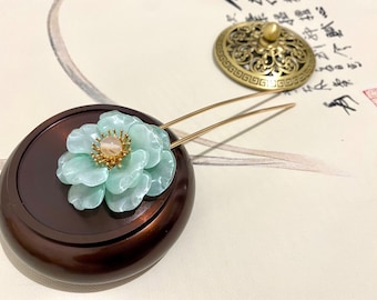 Puede incluir: Una horquilla para el pelo con forma de flor verde claro con un centro dorado y dos puntas doradas. La horquilla está apoyada sobre una superficie de madera marrón oscuro.