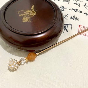 May include: A gold hair stick with a pearl and crystal bead, a brown wooden box with a gold inscription, and a white background with black calligraphy.