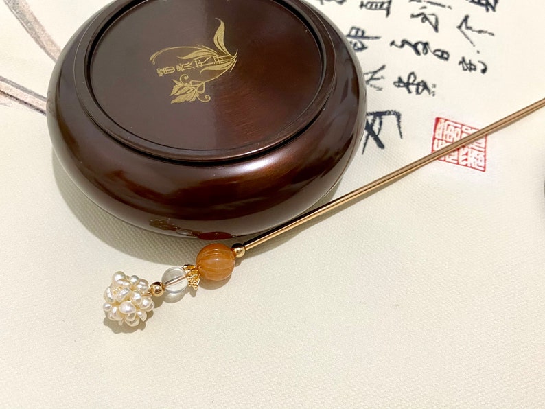 May include: A gold hair stick with a pearl and crystal bead, a brown wooden box with a gold inscription, and a white background with black calligraphy.