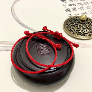 May include: Two red string bracelets with knots on a dark brown wooden surface. The bracelets are tied together and are on a round wooden surface.