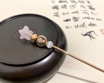 May include: A decorative hair stick with a star-shaped, light pink crystal, gold beads, and a gold-colored metal shaft. The hair accessory is resting on a dark brown surface, with calligraphy in the background.