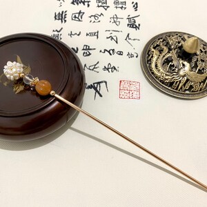 May include: A gold hair stick with a pearl and orange stone embellishment. The stick is lying on a wooden surface next to a small, round, brass incense burner with a dragon and phoenix design.