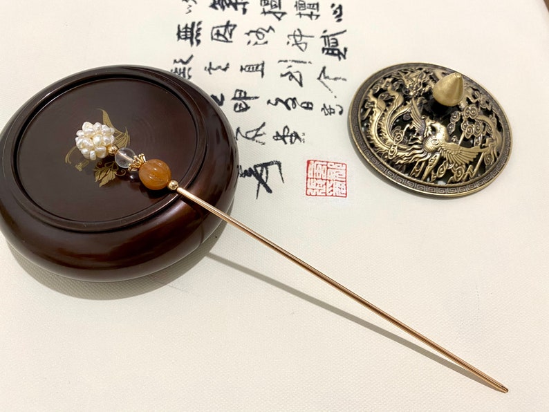 May include: A gold hair stick with a pearl and orange stone embellishment. The stick is lying on a wooden surface next to a small, round, brass incense burner with a dragon and phoenix design.