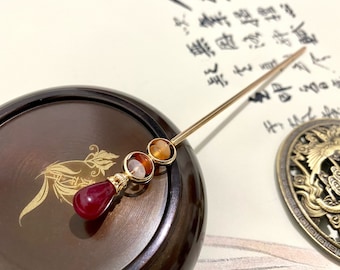 May include: A gold hair stick with three round, colorful beads. The beads are red, orange, and yellow. The stick is resting on a wooden surface.