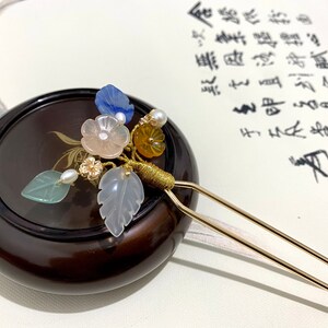 May include: A gold hair stick with a floral design. The design features a pink flower, blue and white leaves, and yellow and white beads.